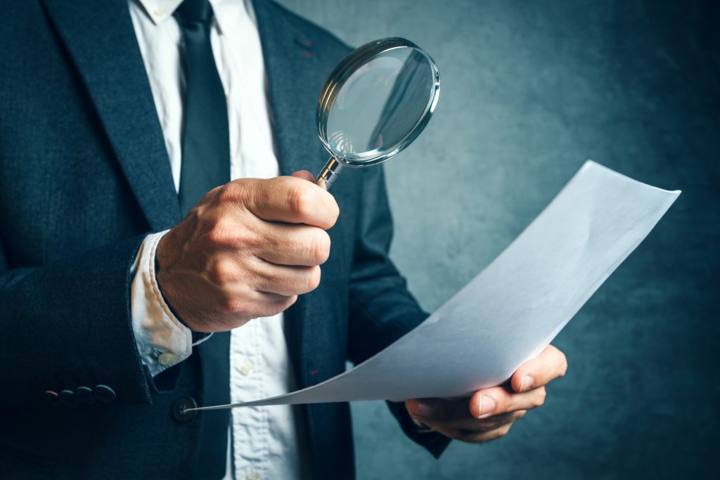 Tax inspector investigating financial documents through magnifying glass, forensic accounting or financial forensics, inspecting offshore company financial papers, documents and reports.